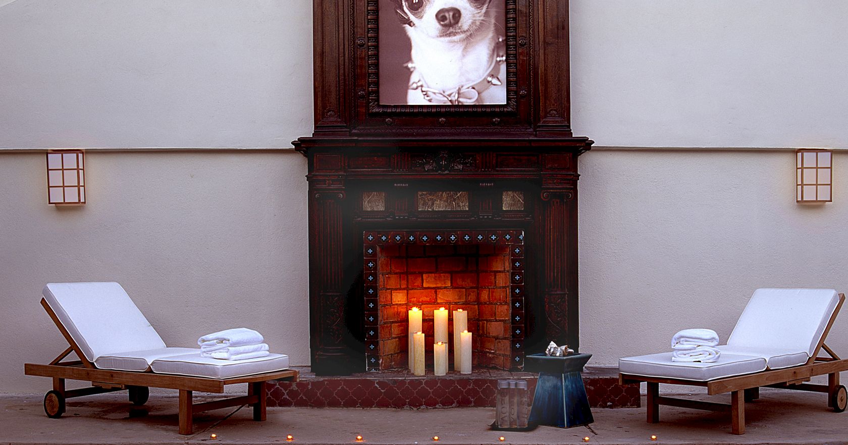 A cozy spa-like patio with two lounge chairs, fluffy towels, and a lit fireplace framed by a creepy portrait of a dog in a dark mantel.