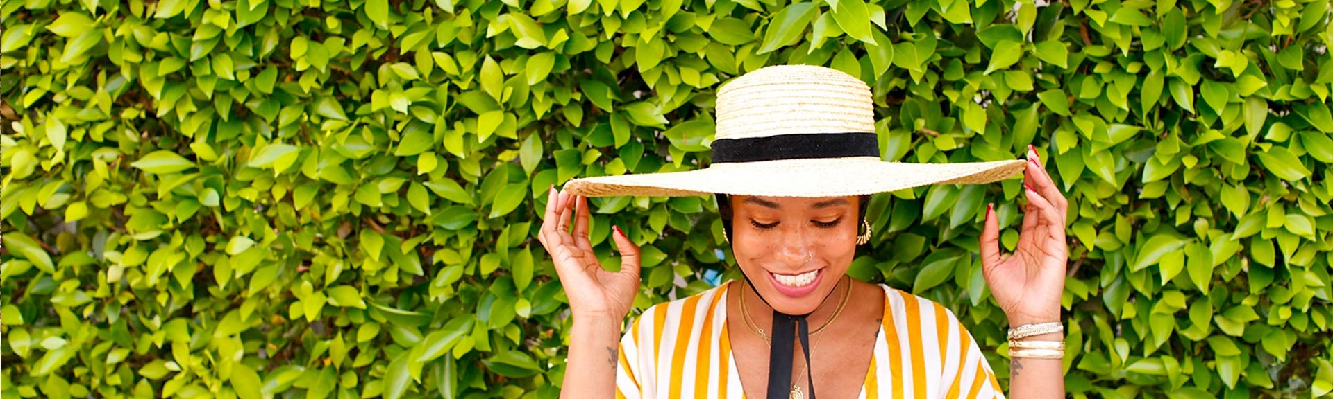 A smiling woman wearing a striped yellow and white outfit and a wide-brimmed hat stands in front of a lush green hedge, holding her hat's brim.