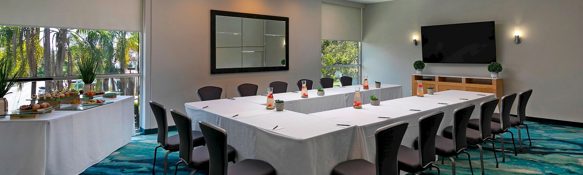 A bright conference room with a U-shaped table setup, white tablecloths, chairs, a wall-mounted TV, snacks on a side table, and large windows letting in natural light.