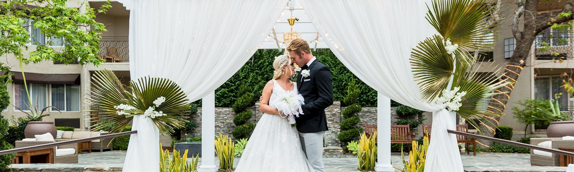 A newlywed couple in a white wedding dress and suit share a kiss under a white draped arch with tropical plants and elegant decor. Ending here.