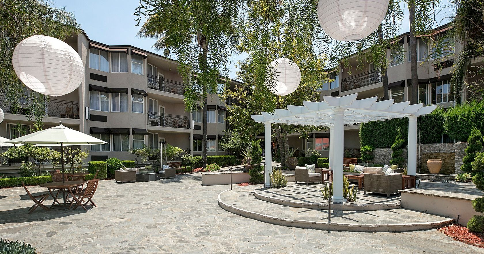 A sunny hotel courtyard with stone paving, round tables under umbrellas, white hanging lanterns, and modern low-rise buildings surrounding a central seating area.