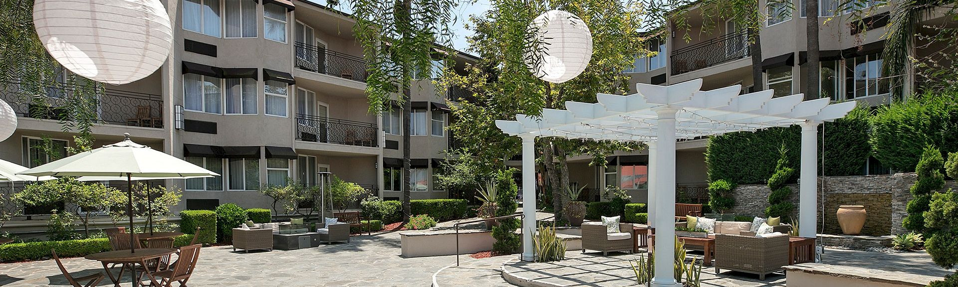 A sunny hotel courtyard with a white pergola, outdoor seating, round paper lanterns, umbrellas, and greenery in a multi-story building backdrop.