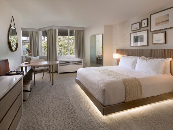 A modern hotel room with a king bed, side lamps, framed art, a desk, a flat-screen TV, and a seating area by large windows.