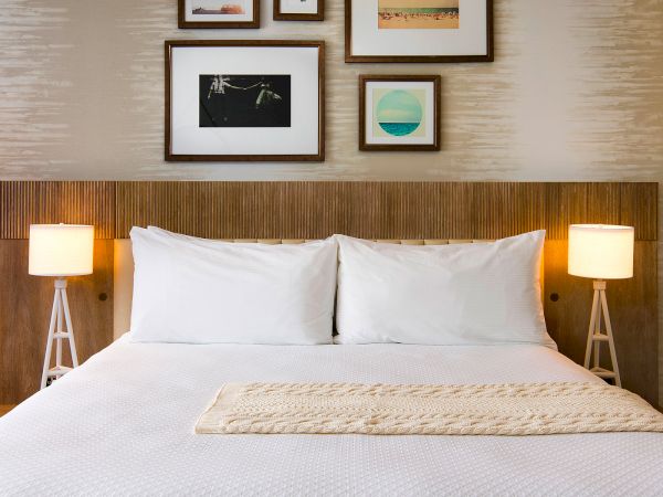 A neatly made hotel-style bedroom with a wooden headboard, two lamps, white bedding, and framed art above the bed, all creating a calm vibe.