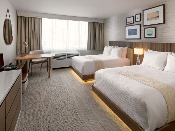 A modern hotel room with two large beds, a desk and chair, a seating area by the window, and framed art above the beds.