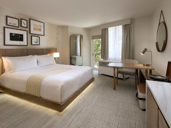 A modern hotel room with a king bed, neutral tones, wall art above the headboard, two bedside lamps, a desk and chair, a TV, and a large window with curtains.