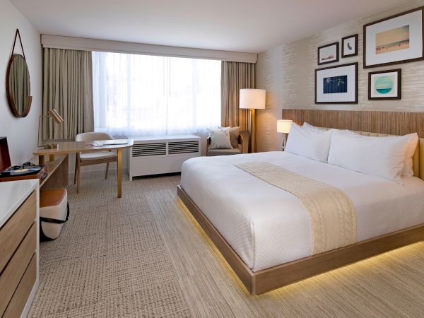 A modern hotel room with a large bed, wooden accents, bedside lamps, a desk, chair, wall art, and a window with sheer curtains.