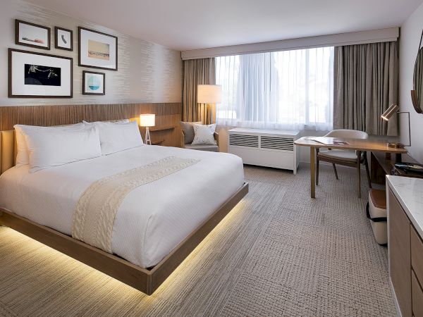 A modern hotel room with a king bed, nightstands, desk, seating area, large window, and beige decor. End.