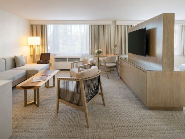A spacious hotel living room with a beige palette, comfortable seating, a wooden coffee table, a wall-mounted TV, and a desk near a large window, all softly lit.