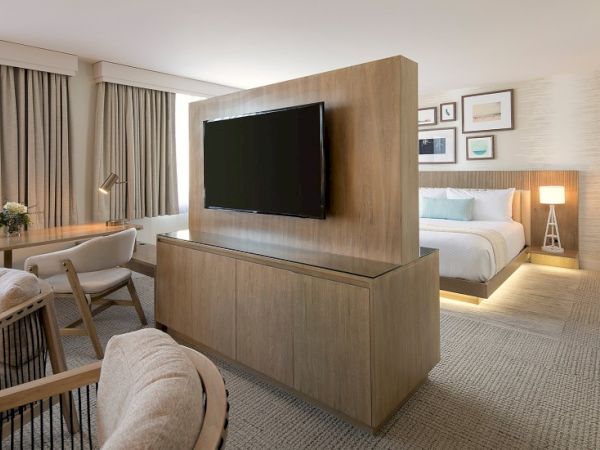 A modern hotel suite with a large wall-mounted TV, wooden partition, seating area, and a cozy bed with warm lighting.