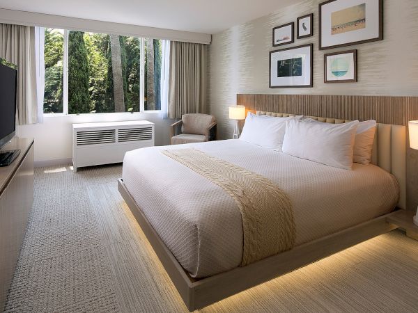 A modern hotel room with a large bed, neutral tones, soft lighting, a wall of framed art, a flat-screen TV, and a window seating area by the window.