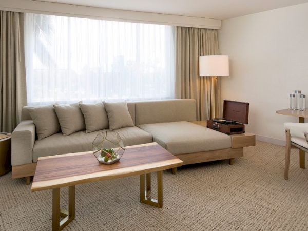 A modern hotel living room: beige sofa, wooden coffee table, neutral tones, large window with sheer curtains, and a tidy seating area.