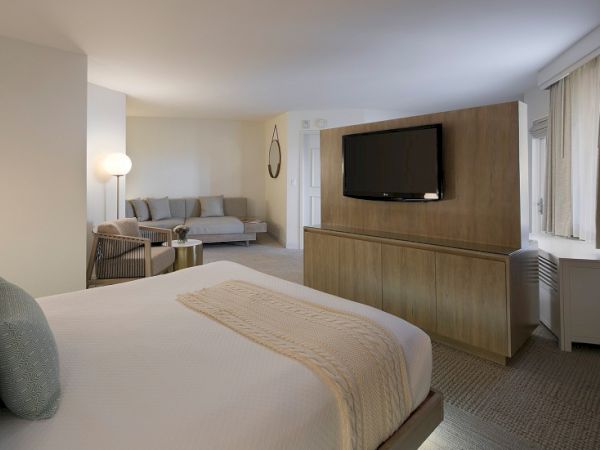 A modern hotel room with a large bed, a seating area, a wall-mounted TV, and soft neutral tones, plus a desk, lamp, and window view.