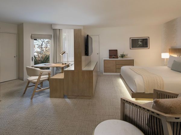 A modern hotel suite with a cozy, open-plan layout: a bed area, a small seating area, a desk, and a light wood divider separating the sleeping space from the living area; neutral tones and soft lighting create a calm ambiance.