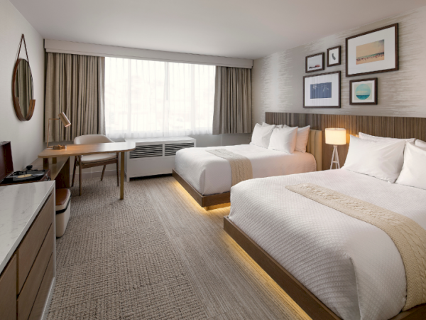 A modern hotel room with two queen beds, a desk, a large window, and warm lighting; neutral decor and framed art above the beds.