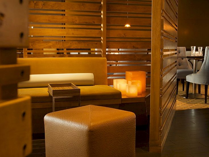 A cozy lounge with warm amber lighting, yellow couches, wooden slat partitions, and a soft ottoman seating area, perfect for relaxing.