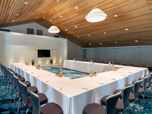 A conference room set for a formal meeting, with a large U-shaped table, white tablecloths, water bottles, and a small pool in the center, under warm wood ceilings.
