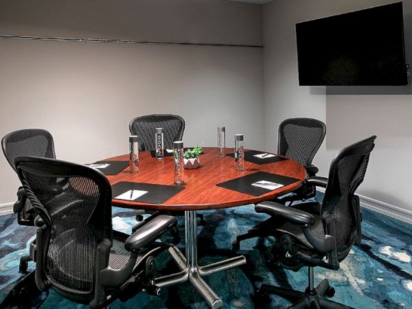 A round conference table with six black ergonomic chairs, papers and water bottles arranged around it, in a modern meeting room with a wall-mounted TV.