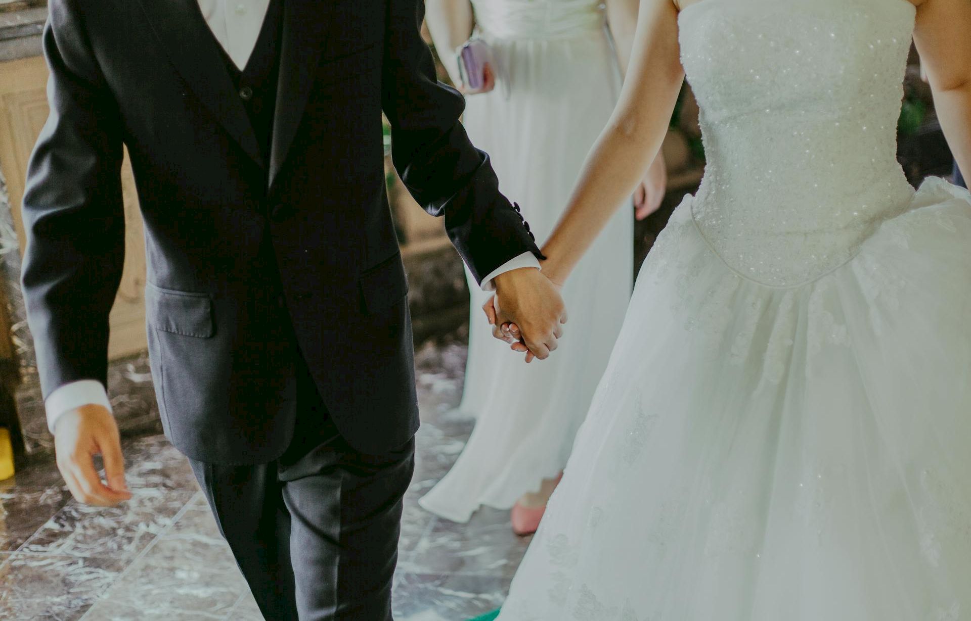 A couple dressed in wedding attire walking hand in hand; a bride in a white gown and a groom in a suit, holding hands.