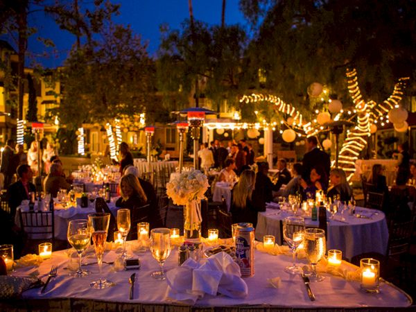 Outdoor evening restaurant scene with lit tables, candles, and people dining under string lights in a festive garden setting.