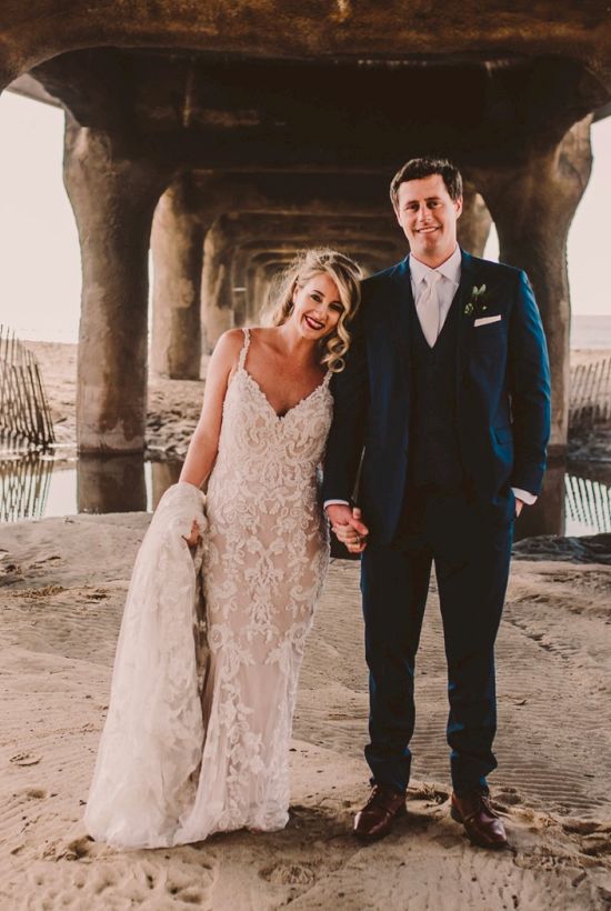 A couple in wedding attire stands hand-in-hand on a sandy beach under a large concrete pier, posing for a photo.