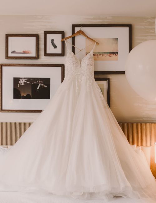 A white wedding dress hangs on a bed&rsquo;s headboard, framed pictures on the wall, and balloons float beside bedside lamps in a cozy room.