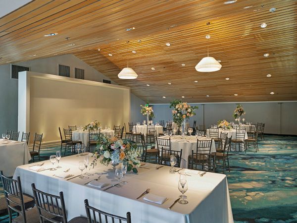 A banquet hall with long white-draped tables, elegant floral centerpieces, black chairs, soft lighting from pendant lamps, and a teal patterned carpet.