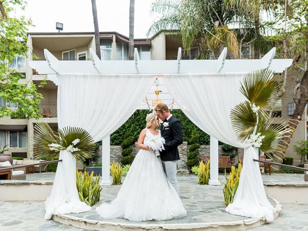 A newlywed couple stands under a white fabric arch, sharing a kiss as guests watch, surrounded by tropical plants and a sunny courtyard.