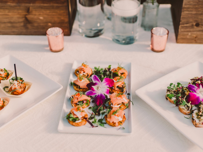 A table of trendy appetizers: bite-size savory bites on white plates, garnished with greens and a bold pink orchid centerpiece.