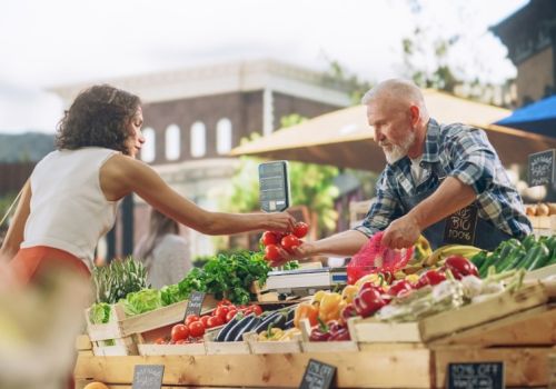 A vendor at a farmers market hands a bag to a shopper amid colorful vegetables and crates under sunny shade.