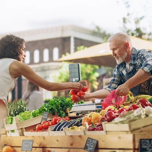 A vendor at a farmers market hands a bag to a shopper amid colorful vegetables and crates under sunny shade.