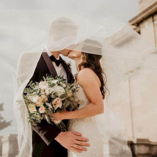 A couple in wedding attire share a kiss under a veil, the bride holding a bouquet as they stand close in front of a grand building.