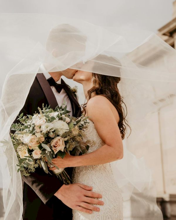 A couple in wedding attire share a kiss under a veil, the bride holding a bouquet as they stand close in front of a grand building.