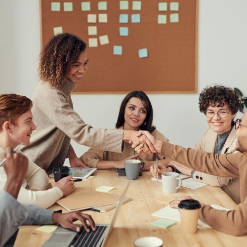 A diverse team sits around a table, shaking hands to celebrate a collaboration while notes and laptops sit nearby.