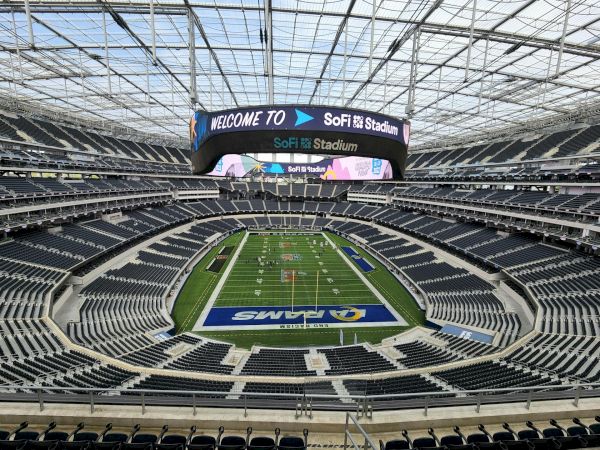 Aerial view of a large football stadium with green field, surrounding stands, and big video board reading &ldquo;WELCOME TO&rdquo; at the top.