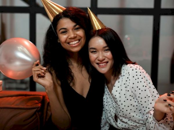 Two smiling women wearing party hats, holding balloons at a celebration, posing closely for a photo.