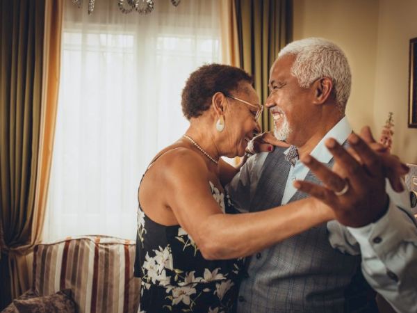 A cheerful elderly couple dances closely in a sunlit living room, smiling and holding hands, sharing a warm, affectionate moment.