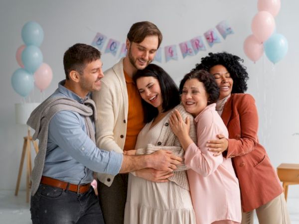 A group of five friends hugs and smiles at a party with pastel balloons and a banner in the background, celebrating together.