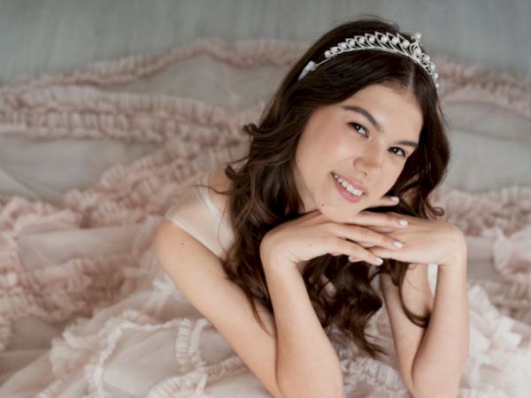 A smiling woman in a white dress with a tiara rests her chin on her hands, lying on a soft, pink, ruffled bedspread.