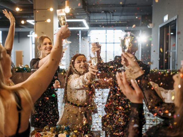 A group of people celebrate indoors, raising trophies and cheering as confetti falls, capturing a joyful party moment.