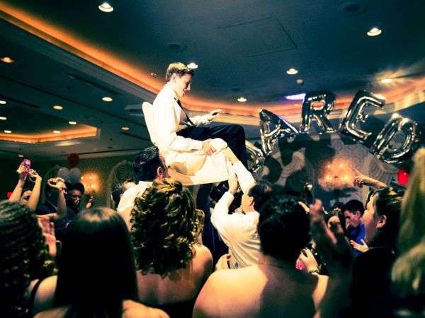 A lively wedding reception with a bride and groom seated on a chair held up by guests, a balloon arch, and a cheering crowd dancing below.