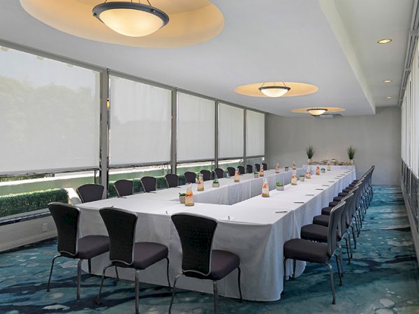 A long conference room with a U-shaped white-table setup, black chairs, water bottles, and large windows with blinds, under ceiling lights.