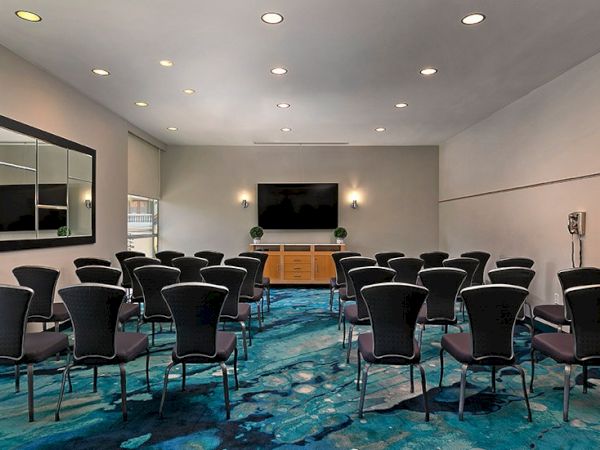 A conference room set up with rows of black chairs facing a wall-mounted screen and a wooden cabinet, blue patterned carpet, and bright overhead lighting.