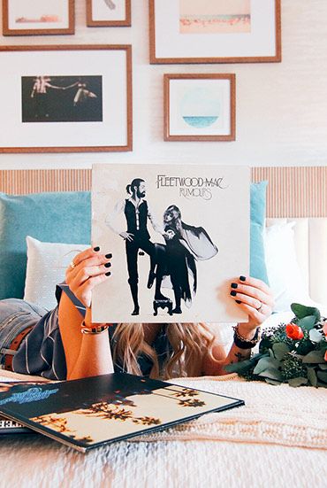 A person lies on a bed holding a black-and-white photo over their face, surrounded by a floral bouquet, magazines, a pillow pile, and wall art above the headboard.