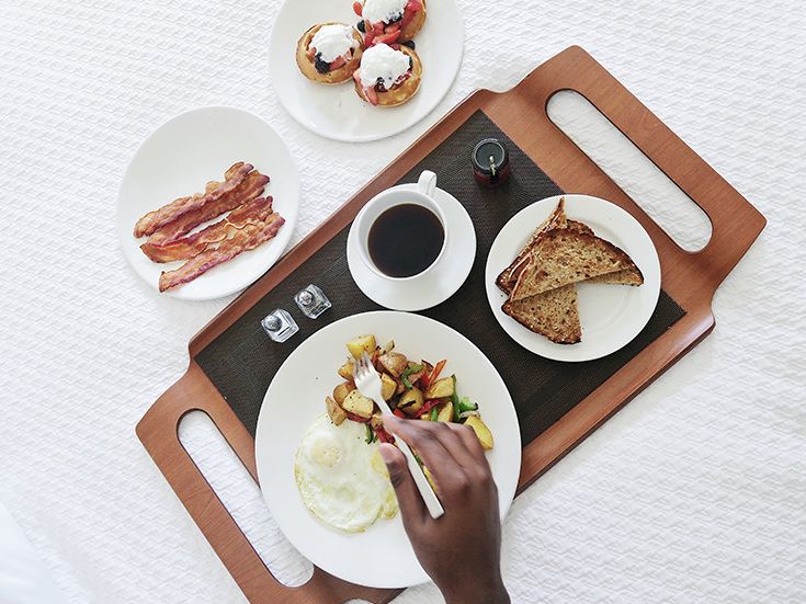 A breakfast scene on a wooden tray: eggs, toast, bacon, fruit salad, coffee, and a hand spreading jam on a sunny-side-up plate, with pastries nearby.