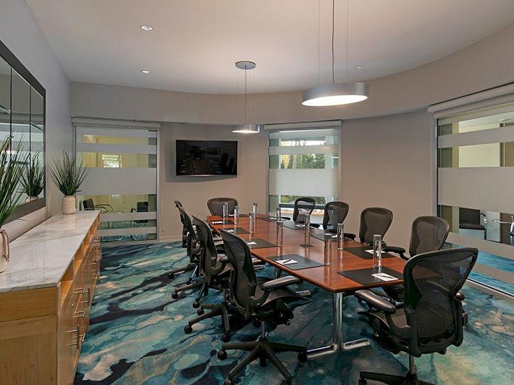 A modern conference room with a long wooden table, black rolling chairs, a wall-mounted TV, glass windows, and a blue patterned carpet, small pendant lights, and plants along a side cabinet. End.