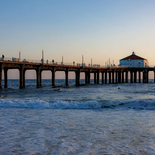 A long pier stretches into the sea with a small building at the end, people walking along the planks as waves roll in under a pastel sunset.