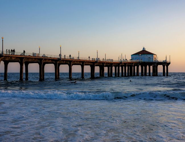 A long pier stretches into the sea with a small building at the end, people walking along the planks as waves roll in under a pastel sunset.