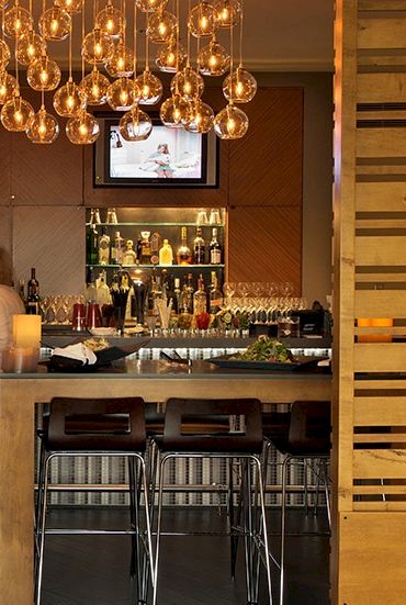 A chic bar interior with warm lighting, people seated at a counter, hanging glass globe decorations, and shelves of bottles behind the bar as TVs show in the background.