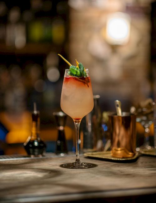 A tropical cocktail in a stemmed glass with a gradient pink to orange hue, garnished with a mint sprig and umbrella, on a bar counter.